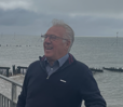 Older man with glasses standing on a pier by a cloudy marina, wearing a dark sweater over a light shirt.