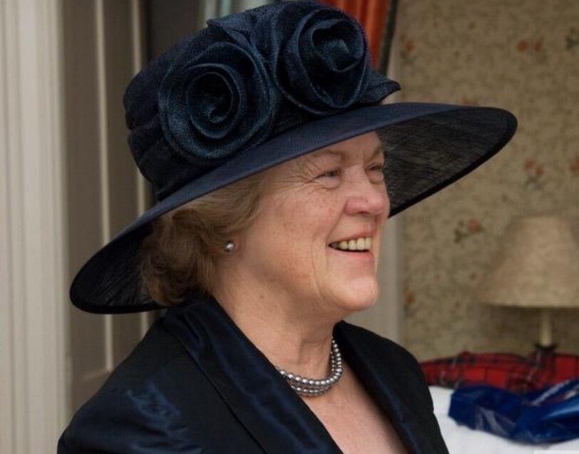 Smiling older woman wearing a navy wide-brim hat with fabric roses, pearl necklace, and dark blazer indoors.