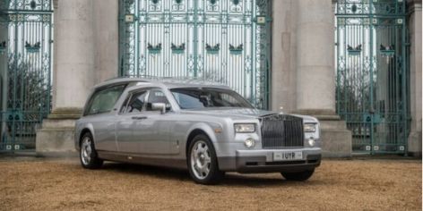 Silver luxury sedan parked on a gravel forecourt in front of a grand teal wrought-iron gate and stone pillars.